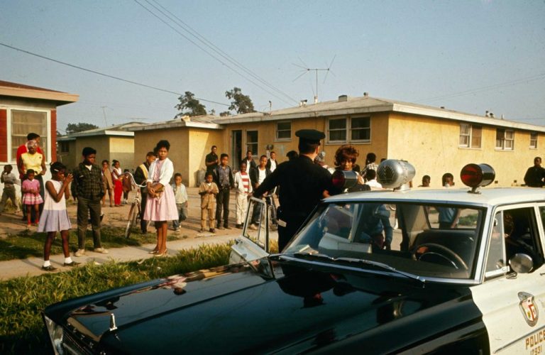 A white police officer stands in the open doorway of a cop car facing a Black woman with curled auburn hair. Over two dozen neighbors, including many young children, stand on the sidewalk in front of yellow one-story homes, observing the police officer and the woman.