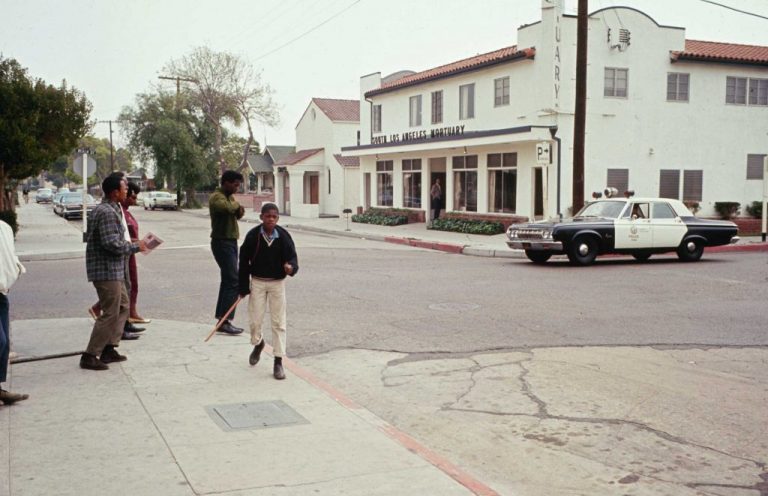 A police car and officer driving it are in front of the South Los Angeles Mortuary. Across the street, six Black commuity members walk towards the street.