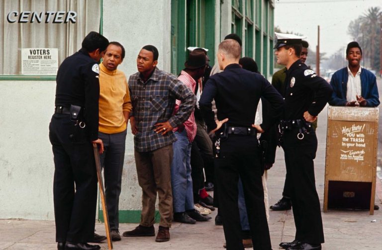 Two Black community members speak with three white police officers on a street corner in front of a green building. Several other community members stand together behind.