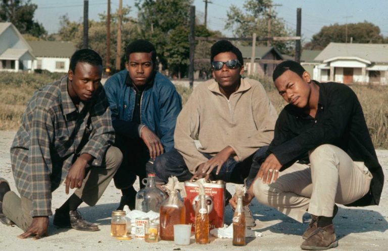 Four young Black activists kneel and look into the camera lens. They pose around an array of molotov cocktails and glassware on the ground.
