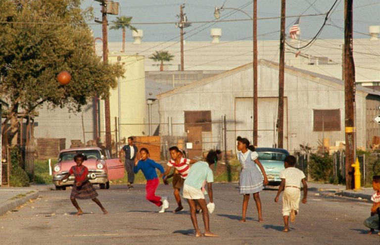 A group of eight young Black children dressed in colorful clothing play ball in the street against the backdrop of industrial buildings. Behind them, a man gets out of a bright pink car.