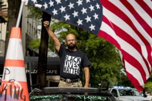 In this June 19, 2021 photo, George Floyd's name is written on the windshield as John Coy wears a shirt that reads Black Lives Matter and stands through his sunroof with his fist in the air at 16th Street Northwest renamed Black Lives Matter Plaza near the White House in Washington.