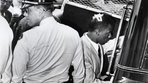 Behind a group of police officers and the door of a large vehicle, John Lewis has a bandage on his scalp and holds a briefcase.